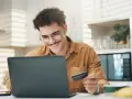 Uśmiechnięty mężczyzna z kartą płatniczą przed laptopem, być może sprawdza limit w koncie przed zakupami online.
