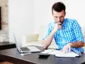 Mężczyzna analizuje dokumenty, zastanawiając się nad prolongatą pożyczki. Na stole laptop, telefon i kalkulator.