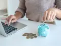 Osoba wkłada monetę do skarbonki, ucząc się, jak oszczędzać pieniądze w domu. Obok leży laptop i garść monet.