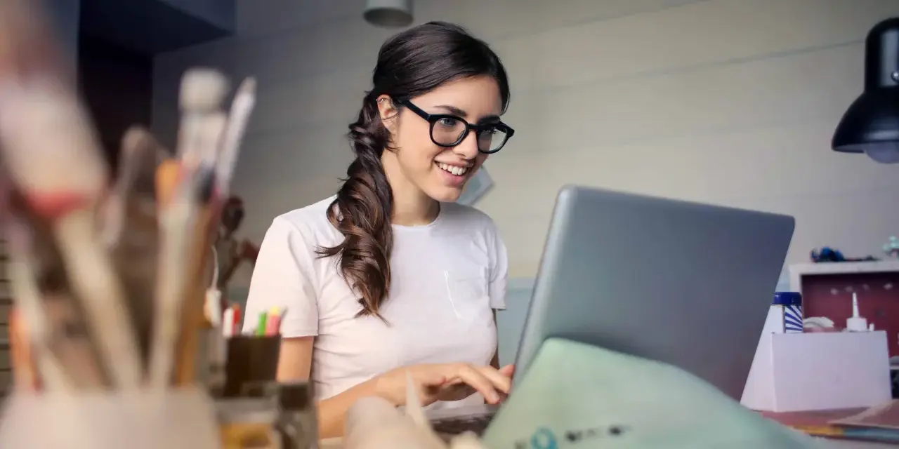 Uśmiechnięta kobieta w okularach pracuje przy laptopie, realizując swoją pracę dodatkową z domu.