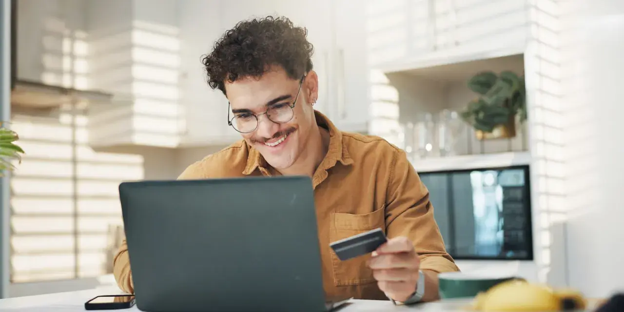 Uśmiechnięty mężczyzna z kartą płatniczą przed laptopem, być może sprawdza limit w koncie przed zakupami online.