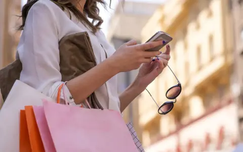 Kobieta z zakupami sprawdza telefon, planując refinansowanie pożyczki.