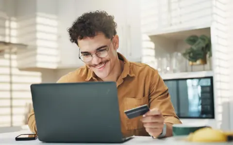 Uśmiechnięty mężczyzna z kartą płatniczą przed laptopem, być może sprawdza limit w koncie przed zakupami online.
