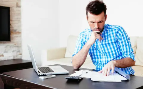 Mężczyzna analizuje dokumenty, zastanawiając się nad prolongatą pożyczki. Na stole laptop, telefon i kalkulator.