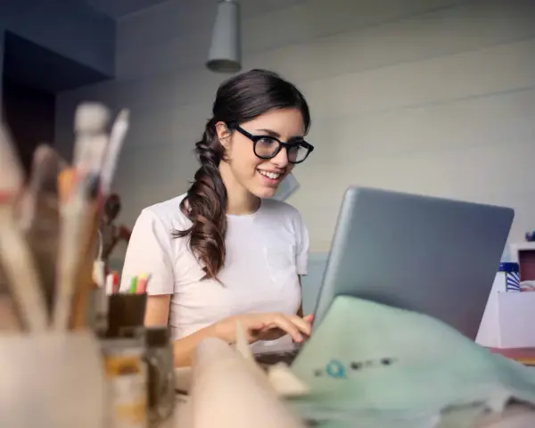 Uśmiechnięta kobieta w okularach pracuje przy laptopie, realizując swoją pracę dodatkową z domu.