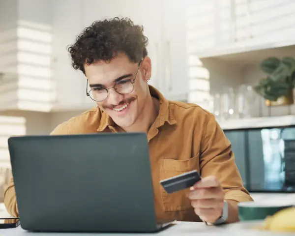 Uśmiechnięty mężczyzna z kartą płatniczą przed laptopem, być może sprawdza limit w koncie przed zakupami online.