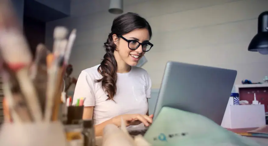 Uśmiechnięta kobieta w okularach pracuje przy laptopie, realizując swoją pracę dodatkową z domu.