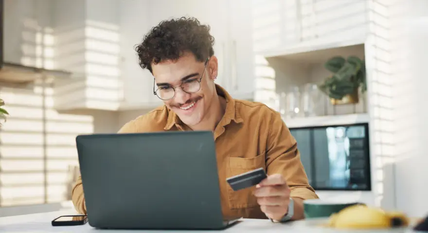 Uśmiechnięty mężczyzna z kartą płatniczą przed laptopem, być może sprawdza limit w koncie przed zakupami online.