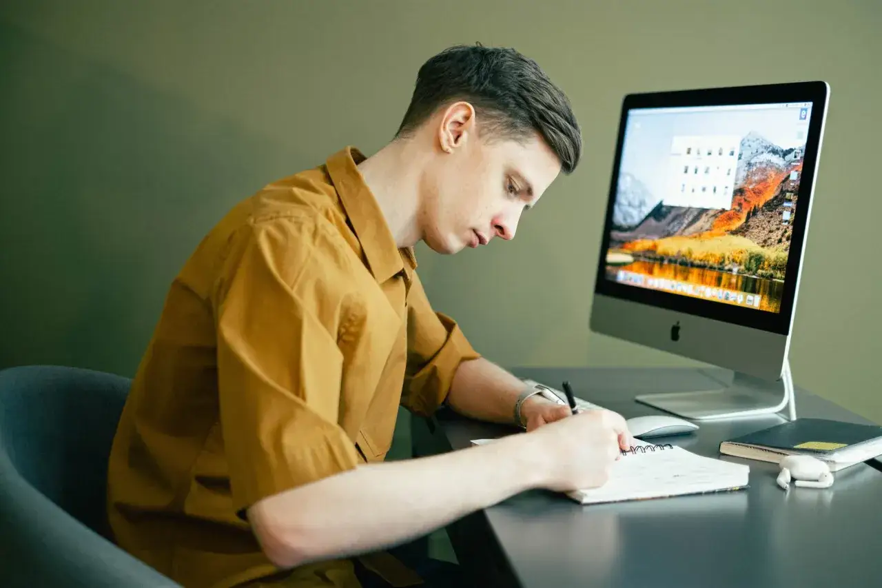 Młody mężczyzna siedzi przy biurku, notując coś w zeszycie. Obok stoi komputer. Zastanawia się, jak zarobić 100 zł dziennie przez internet.