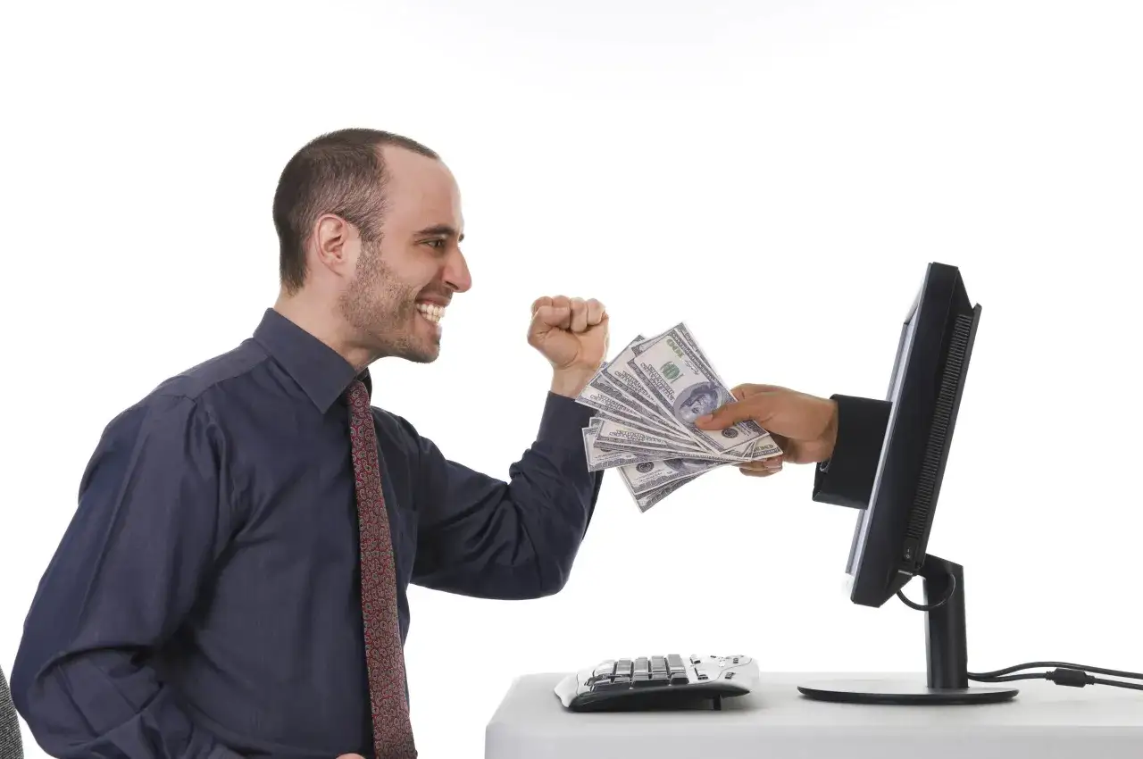 Uśmiechnięty mężczyzna otrzymuje stos banknotów z monitora komputera. To symbol zarabiania na klikaniu w internecie.